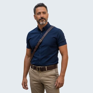 May include: A man in a navy blue short-sleeved shirt, khaki trousers, and a brown leather belt with a gold buckle. A brown leather messenger bag is worn over the shoulder.