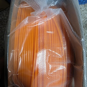 May include: A spool of orange filament for 3D printing, wrapped in clear plastic packaging.