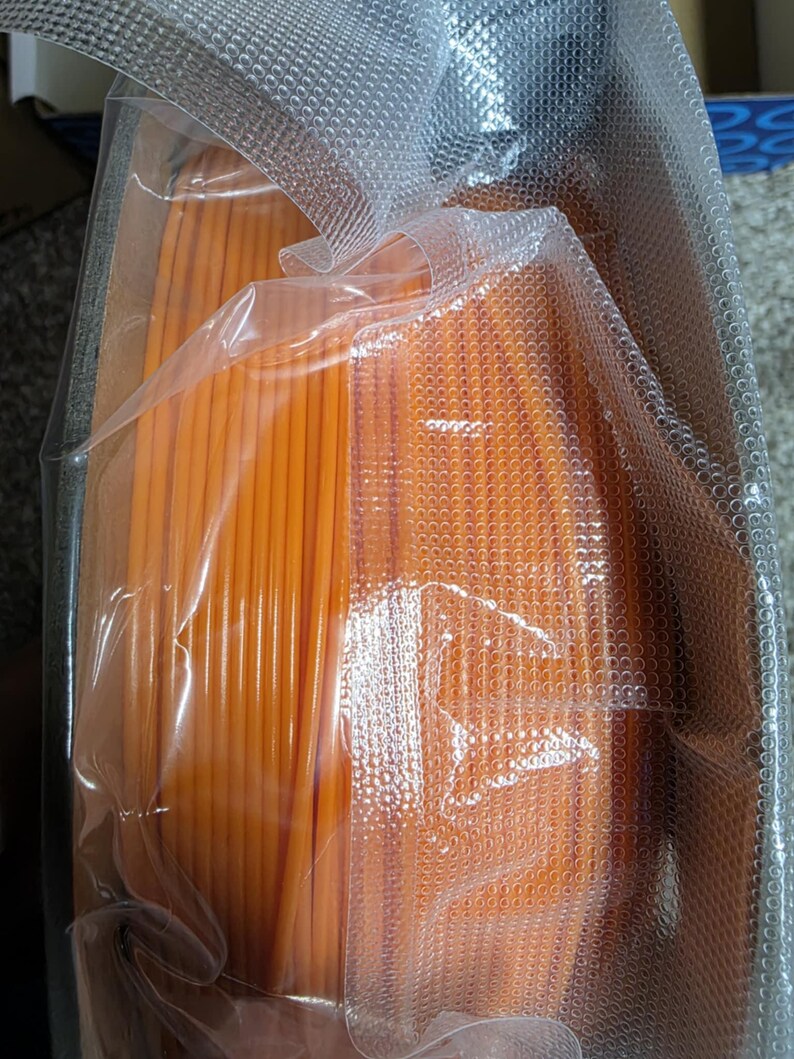 May include: A spool of orange filament for 3D printing, wrapped in clear plastic packaging.