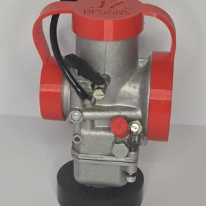 Puede incluir: Un carburador plateado con detalles en rojo y el texto "JZ DESIGNS" en la parte superior. El carburador tiene una l&iacute;nea de combustible negra y est&aacute; montado sobre una base negra.