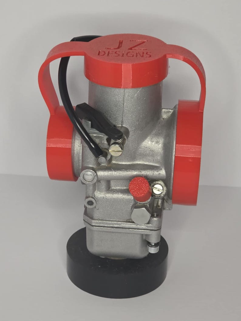 Puede incluir: Un carburador plateado con detalles en rojo y el texto "JZ DESIGNS" en la parte superior. El carburador tiene una l&iacute;nea de combustible negra y est&aacute; montado sobre una base negra.