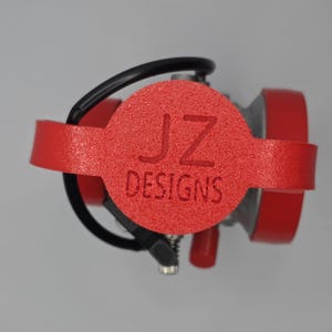 Puede incluir: Un objeto rojo texturizado con el texto "JZ DESIGNS" se muestra. Tiene un centro circular y dos piezas laterales rojas. Tambi&eacute;n se ven un cable negro y componentes plateados.