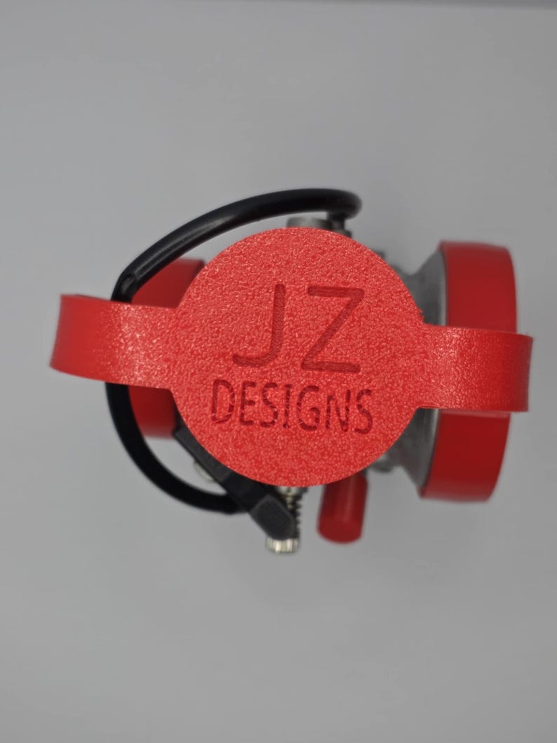 Puede incluir: Un objeto rojo texturizado con el texto "JZ DESIGNS" se muestra. Tiene un centro circular y dos piezas laterales rojas. Tambi&eacute;n se ven un cable negro y componentes plateados.