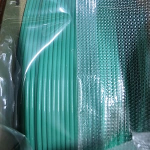 May include: A close-up of a spool of green filament for a 3D printer. The filament is wrapped in clear plastic and is sitting on a white surface.