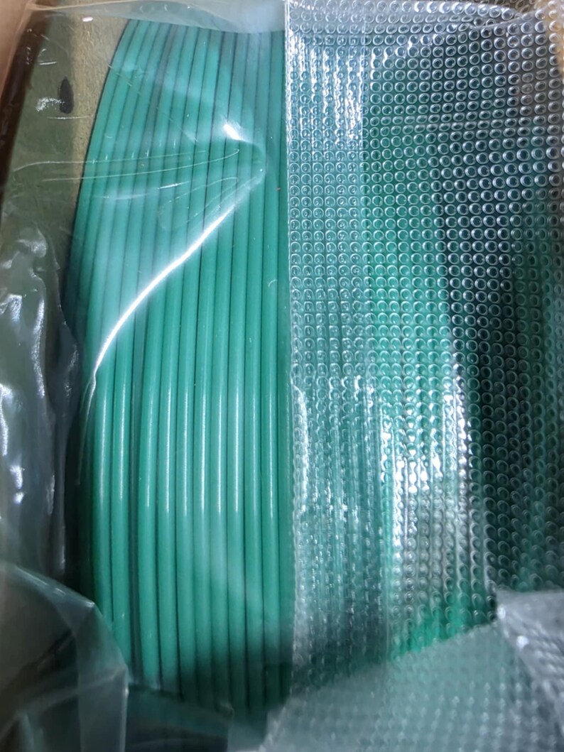 May include: A close-up of a spool of green filament for a 3D printer. The filament is wrapped in clear plastic and is sitting on a white surface.