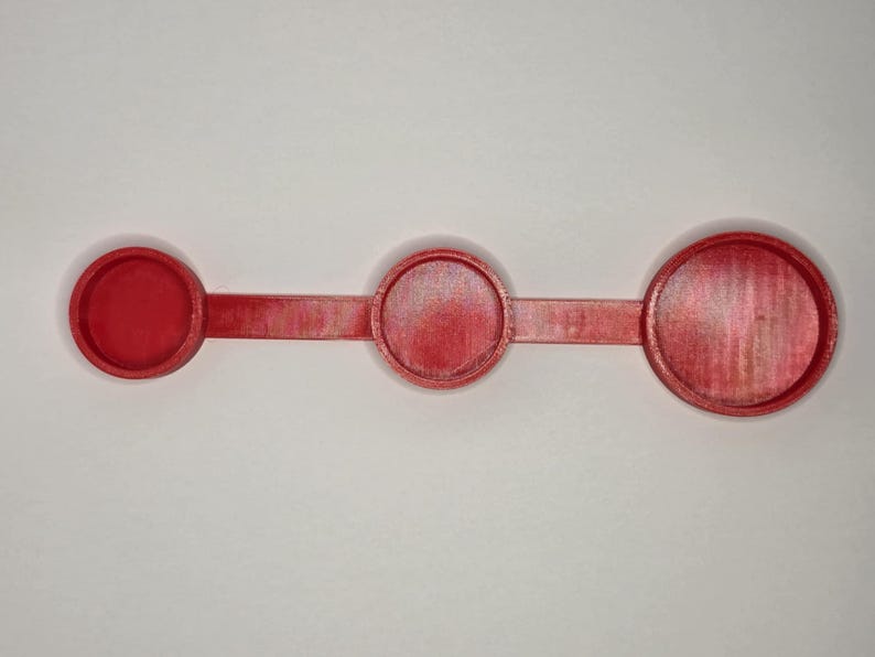 Puede incluir: Un juego de cucharas medidoras de pl&aacute;stico rojo. Cuenta con tres tazas medidoras circulares de diferentes tama&ntilde;os, conectadas por un mango plano y delgado. El juego est&aacute; sobre un fondo blanco.