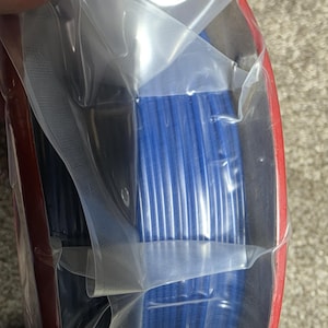 May include: A spool of blue filament for 3D printing is wrapped in clear plastic and packaged in a red box.