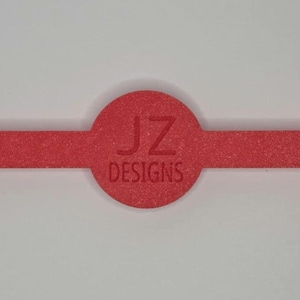 Puede incluir: Un elemento de dise&ntilde;o rojo texturizado, horizontal, con tres componentes circulares conectados por una barra rectangular estrecha. El c&iacute;rculo central presenta el texto "JZ DESIGNS" en rojo m&aacute;s oscuro. El dise&ntilde;o est&aacute; sobre un fondo blanco.