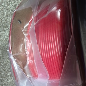 May include: A spool of red filament for a 3D printer, wrapped in clear plastic packaging.