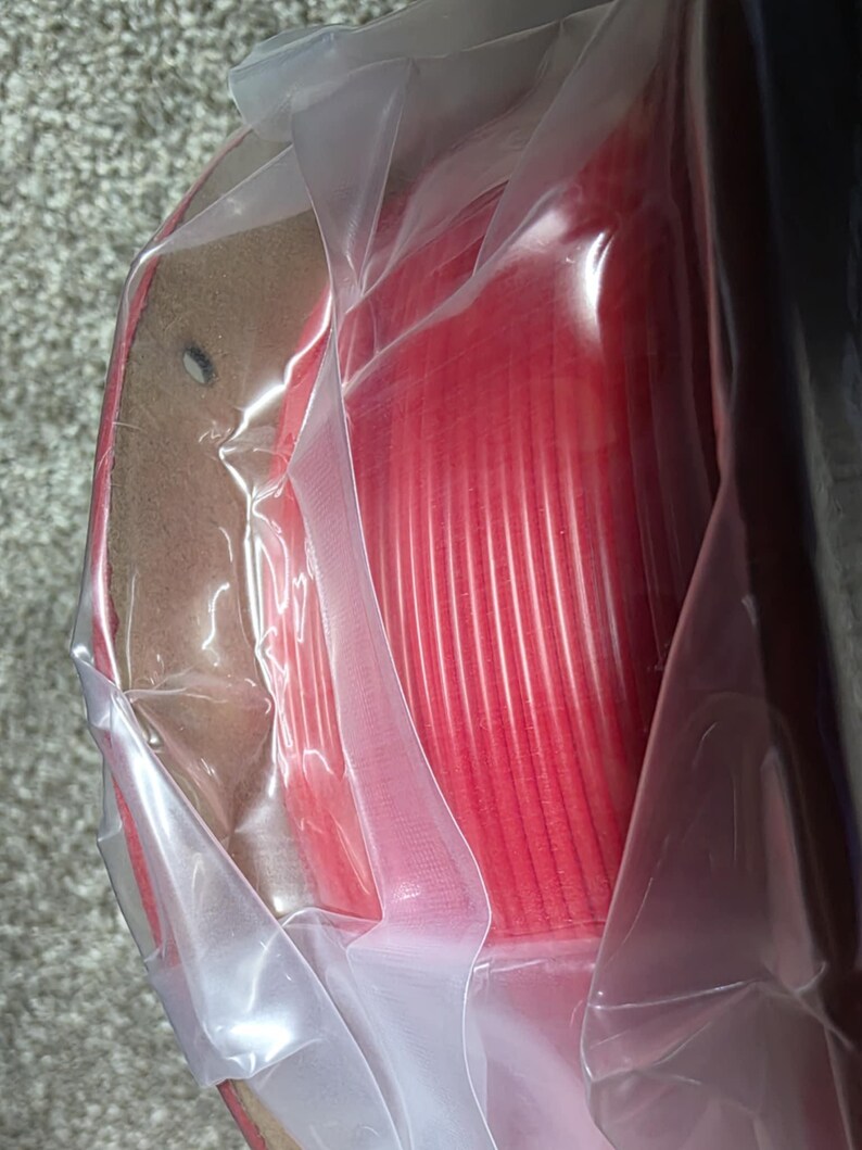 May include: A spool of red filament for a 3D printer, wrapped in clear plastic packaging.