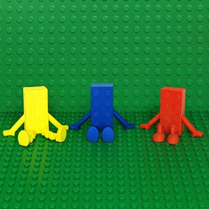 May include: Three colorful Lego-style figures: yellow, blue, and red, each with arms and legs. They are arranged on a green Lego baseplate. The figures are in a sitting position.
