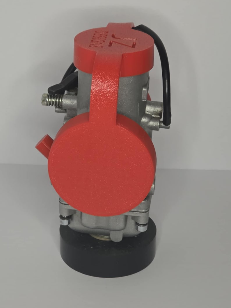 Puede incluir: Un componente mec&aacute;nico plateado con una cubierta roja. La cubierta tiene el texto blanco "DESIGNS" y "7L". Tambi&eacute;n se ven un cable negro y una base negra.