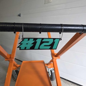 May include: A teal blue and black plastic sign with the number #121 hanging from a black metal bar.
