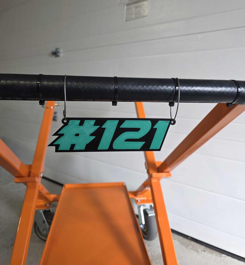 May include: A teal blue and black plastic sign with the number #121 hanging from a black metal bar.