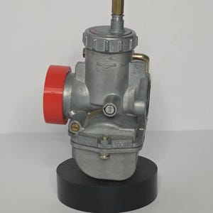 Puede incluir: Un carburador plateado con un componente rojo y un racor de lat&oacute;n. El carburador est&aacute; montado sobre una base negra. El n&uacute;mero "590890" es visible en el lateral.