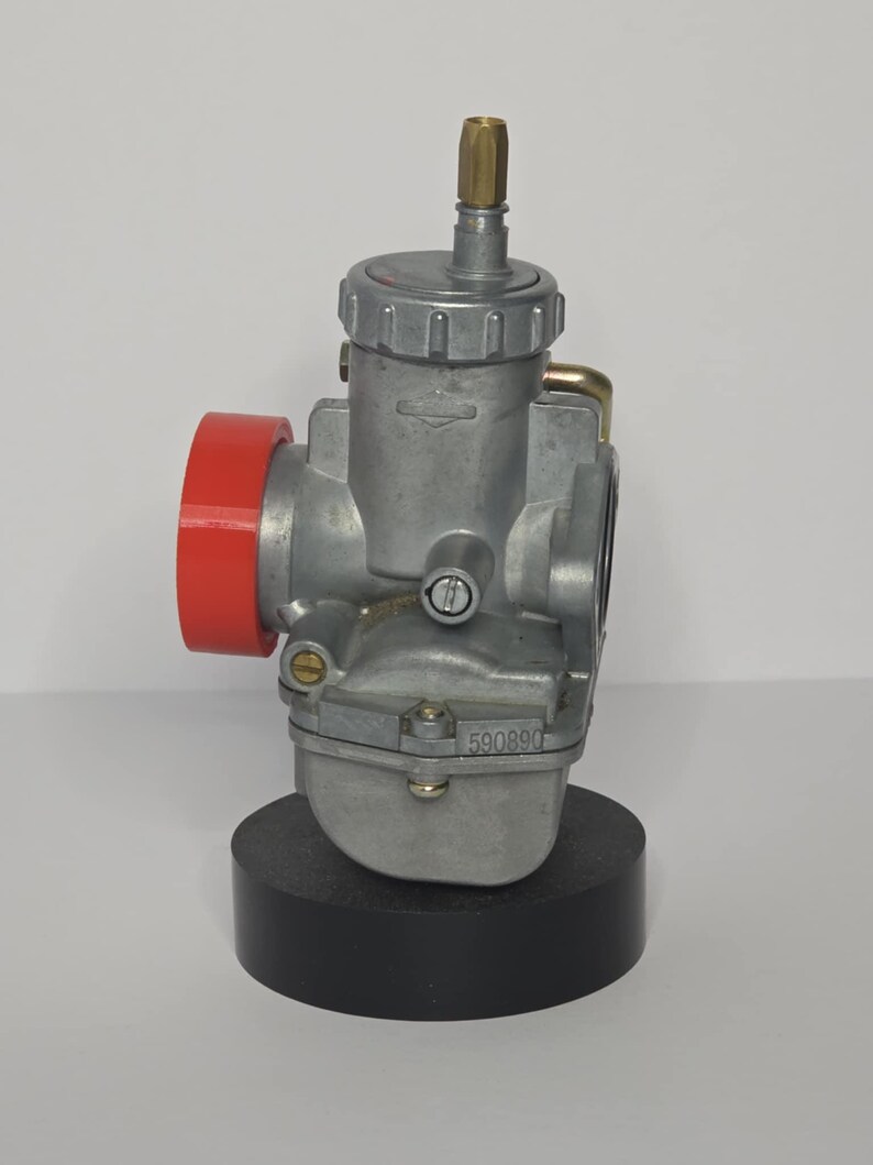Puede incluir: Un carburador plateado con un componente rojo y un racor de lat&oacute;n. El carburador est&aacute; montado sobre una base negra. El n&uacute;mero "590890" es visible en el lateral.
