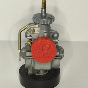 Puede incluir: Un carburador plateado con una tapa roja que dice "JZ DESIGNS". El carburador tiene un accesorio de lat&oacute;n y est&aacute; montado sobre una base negra. El fondo es blanco liso.