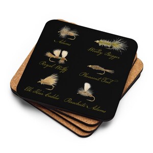 May include: A set of square coasters with a black surface, displaying illustrations of fishing flies and their names in gold script. The coasters are stacked on a cork base.