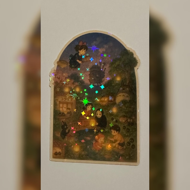Op de afbeelding: Een holografische sticker met een sc&egrave;ne uit een Japanse animatiefilm. De afbeelding toont personages in een bos met gloeiende sterren en lantaarns. De sticker heeft een kenmerkende, boogvormige vorm.