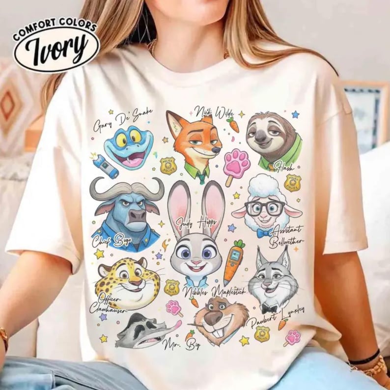 May include: Ivory-colored t-shirt featuring cartoon characters from a movie. The design includes characters like Judy Hopps, Nick Wilde, and Flash, with their names written below. The shirt has the Comfort Colors logo in the upper left corner.