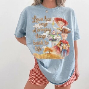 May include: A light blue t-shirt featuring a graphic print of Woody, Buzz Lightyear, and Jessie from Toy Story. The text reads, Love has ways of bringing things back to life, set against a background of clouds and a tree on a golden hill.