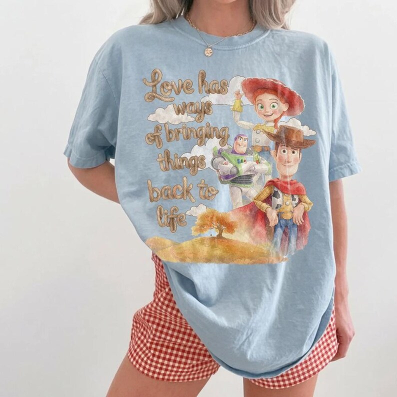 May include: A light blue t-shirt featuring a graphic print of Woody, Buzz Lightyear, and Jessie from Toy Story. The text reads, Love has ways of bringing things back to life, set against a background of clouds and a tree on a golden hill.