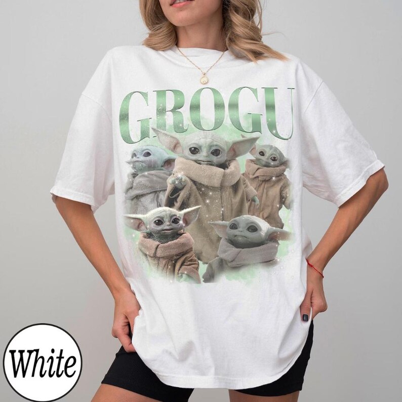 May include: A white short-sleeve t-shirt featuring a graphic print of five Grogu characters in tan robes. The text GROGU is printed in large green letters above the characters. The design includes subtle sparkling accents on a light green background.