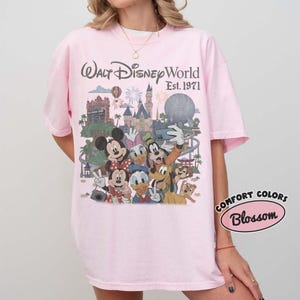 May include: A light pink t-shirt featuring a graphic print of Walt Disney World Est. 1971 with various cartoon characters including Mickey Mouse, Minnie Mouse, Donald Duck, Daisy Duck, Goofy, and Pluto in front of the park landmarks.