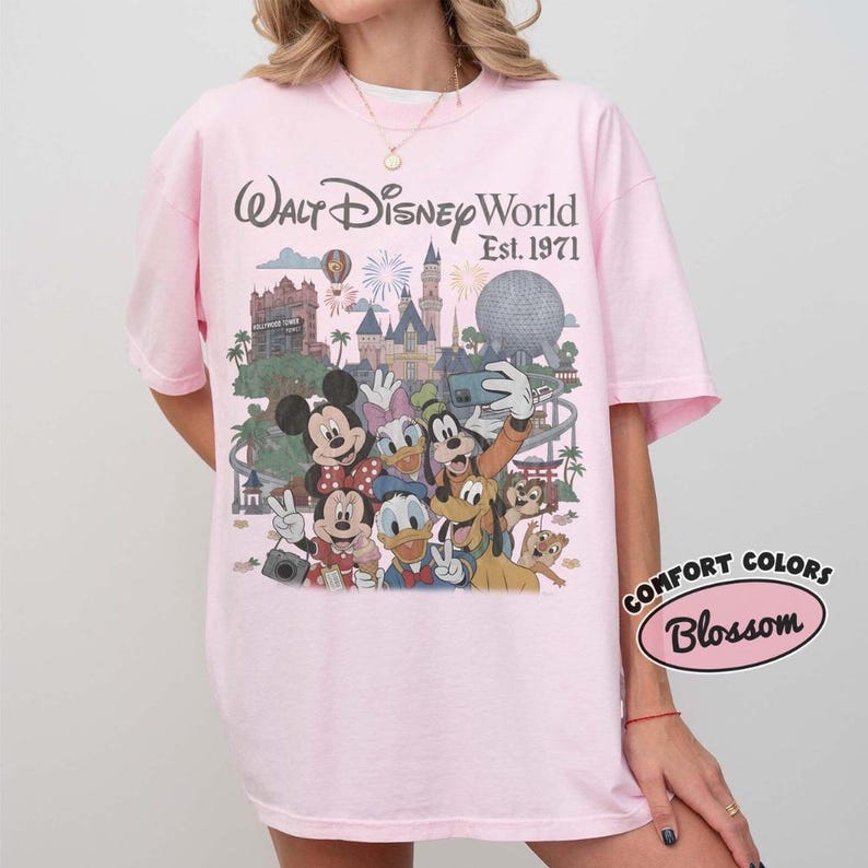 May include: A light pink t-shirt featuring a graphic print of Walt Disney World Est. 1971 with various cartoon characters including Mickey Mouse, Minnie Mouse, Donald Duck, Daisy Duck, Goofy, and Pluto in front of the park landmarks.