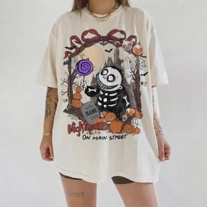 Może przedstawiać: Kremowy t-shirt z grafiką przedstawiającą szkielet trzymający fioletowego lizaka. Wz&oacute;r zawiera nagrobek z napisem "NAME 2026", dynie, zamek oraz czerwono-czarny napis "Nightmare ON MAIN STREET" umieszczony na środku koszulki.