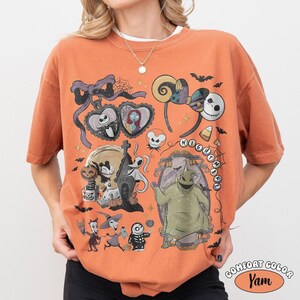 Op de afbeelding: Een oranje t-shirt met een grafische print van personages en motieven uit The Nightmare Before Christmas. Het ontwerp bevat Jack Skellington, Sally, Zero, Oogie Boogie en diverse thematische iconen zoals muisoren, strikken en spinnenwebben.