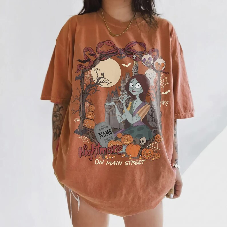 Może przedstawiać: Pomarańczowy t-shirt z kr&oacute;tkim rękawem z nadrukiem graficznym przedstawiającym postać kobiecą z długimi ciemnymi włosami, zamek, dynie i nagrobek z napisem NAME 2026, dużą kokardę, nietoperze oraz napis Nightmare ON MAIN STREET.