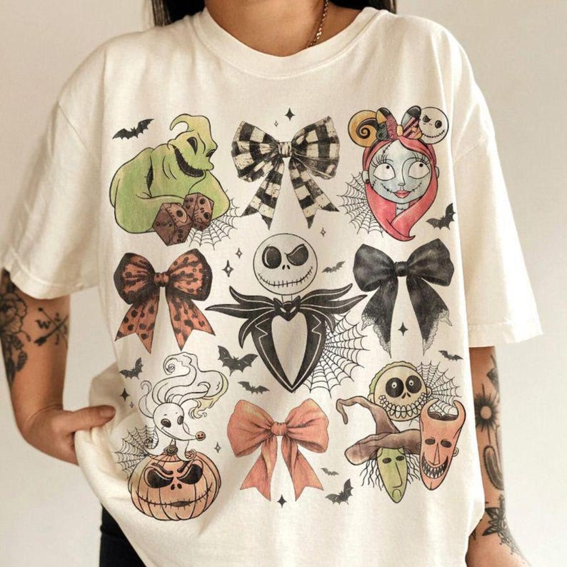 Puede incluir: Una camiseta de manga corta color hueso con un estampado gr&aacute;fico de varios personajes y lazos. El dise&ntilde;o incluye una figura verde con dados, una cara de calavera, un perro fantasma sobre una calabaza y varios lazos en negro, cuadros y rosa.