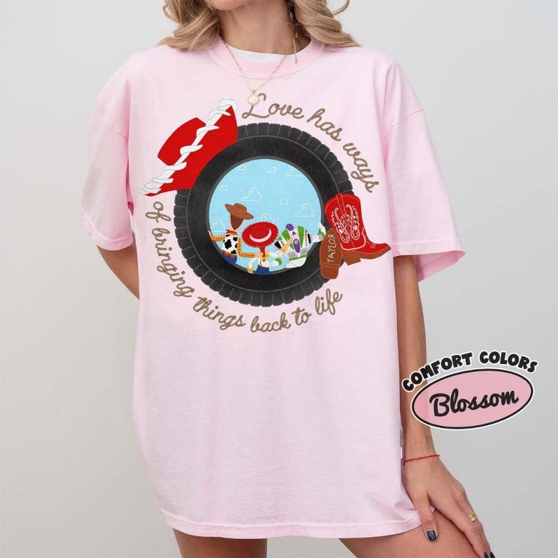 May include: A light pink t-shirt featuring a graphic of a black tire surrounding a blue sky scene with cartoon characters. The text reads Love has ways of bringing things back to life. A red cowboy hat and a red cowboy boot are positioned on the tire.