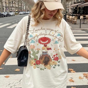 May include: An off-white short-sleeve t-shirt featuring a graphic of a cowgirl on a horse surrounded by cacti and flowers. The text reads, Love has ways of bringing things back to life, accompanied by musical notes and small blue clouds.
