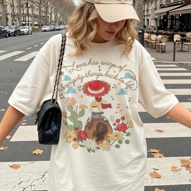 May include: An off-white short-sleeve t-shirt featuring a graphic of a cowgirl on a horse surrounded by cacti and flowers. The text reads, Love has ways of bringing things back to life, accompanied by musical notes and small blue clouds.