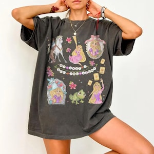 May include: Dark gray t-shirt featuring a colorful graphic design with the text "At Last I See The Light". The design includes illustrations of a horse, a princess, a frog, and floating lanterns. The shirt has a vintage, worn look.