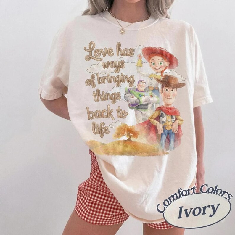 May include: An ivory short-sleeve t-shirt featuring a graphic print of characters from Toy Story, including Woody, Buzz Lightyear, and Jessie. The text reads, Love has ways of bringing things back to life. The shirt has a crew neckline and loose fit.