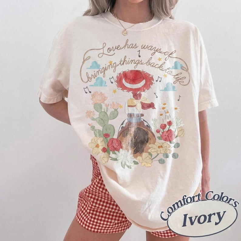 May include: An ivory t-shirt featuring a graphic of a cowgirl in a red hat riding a horse, surrounded by cacti and flowers. The text reads, Love has ways of bringing things back to life, accompanied by musical notes and small blue clouds.