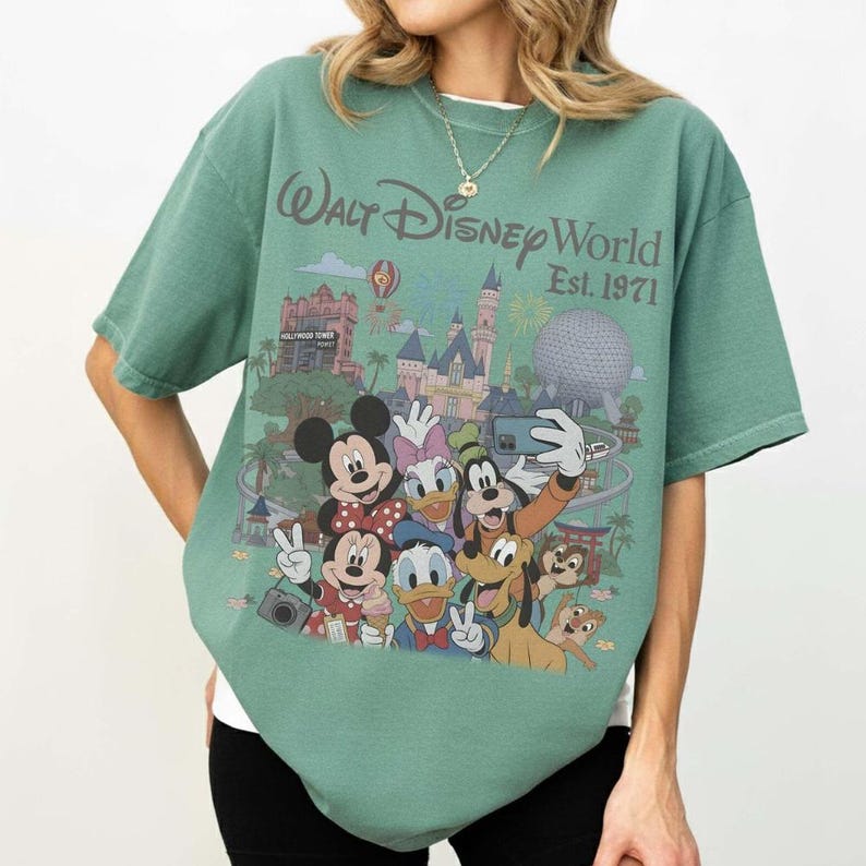May include: A sage green short-sleeve t-shirt featuring a graphic print of Walt Disney World Est. 1971. The design includes illustrations of Mickey Mouse, Minnie Mouse, Donald Duck, Daisy Duck, Goofy, Pluto, Chip, and Dale taking a selfie.