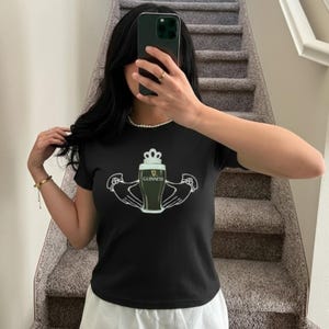 May include: Black t-shirt with a Guinness design. The graphic features a pint of Guinness, a crown, and hands. The short-sleeved shirt is made of a soft material. The shirt is worn by a person on a staircase.