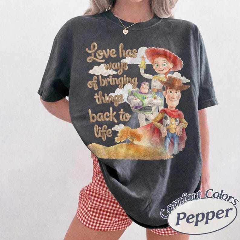 May include: A charcoal gray short-sleeve t-shirt featuring a graphic print of Buzz Lightyear, Jessie, and Woody from Toy Story. The text reads, Love has ways of bringing things back to life. The shirt has a crew neckline and a relaxed, loose fit.