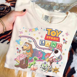 Pode incluir: Camisola creme com um gr&aacute;fico colorido "Toy Story Land". O design inclui personagens como Slinky Dog, Buzz Lightyear e Woody, juntamente com foguetes, flores e o logotipo Toy Story. A camisola tem gola redonda e mangas compridas.