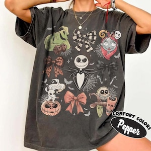 Puede incluir: Una camiseta gris carb&oacute;n con un estampado gr&aacute;fico de personajes de Pesadilla antes de Navidad, incluyendo a Jack Skellington, Sally, Oogie Boogie y Zero, intercalados con varios lazos decorativos, telara&ntilde;as y peque&ntilde;os murci&eacute;lagos.