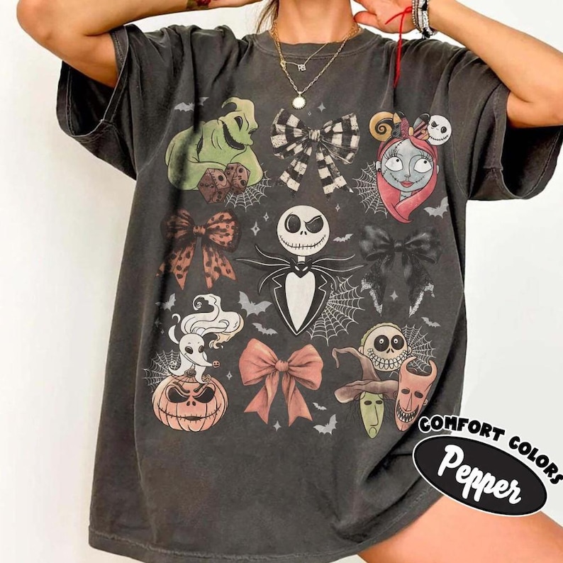 Puede incluir: Una camiseta gris carb&oacute;n con un estampado gr&aacute;fico de personajes de Pesadilla antes de Navidad, incluyendo a Jack Skellington, Sally, Oogie Boogie y Zero, intercalados con varios lazos decorativos, telara&ntilde;as y peque&ntilde;os murci&eacute;lagos.