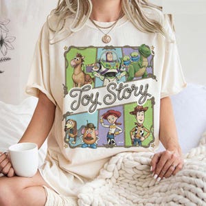 Può includere: T-shirt color crema con una grafica colorata dei personaggi del film d'animazione "Toy Story". Il design include Woody, Buzz Lightyear e altri personaggi, con le parole "Toy Story" in un carattere decorativo.