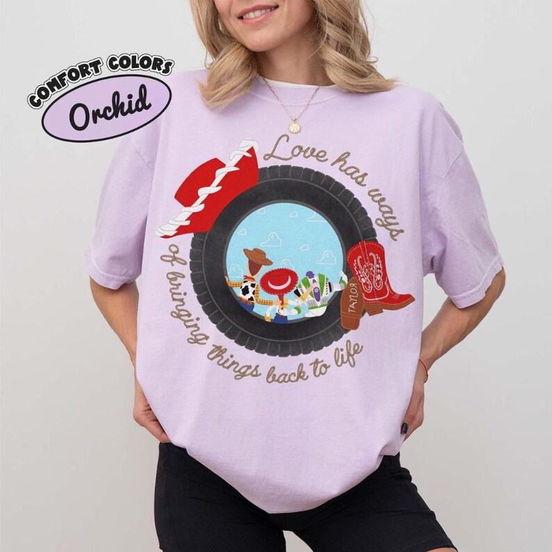 May include: A light purple t-shirt featuring a graphic of a black tire framing cartoon characters. The text reads, Love has ways of bringing things back to life. A red cowboy hat and a red cowboy boot are positioned on the tire. The shirt is labeled Orchid.