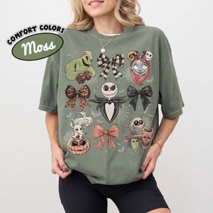 Puede incluir: Una camiseta verde musgo con un estampado gr&aacute;fico de personajes y lazos de Pesadilla antes de Navidad, incluyendo a Jack Skellington, Sally, Zero y Oogie Boogie, dispuestos con detalles de telara&ntilde;as y peque&ntilde;os murci&eacute;lagos en la tela.