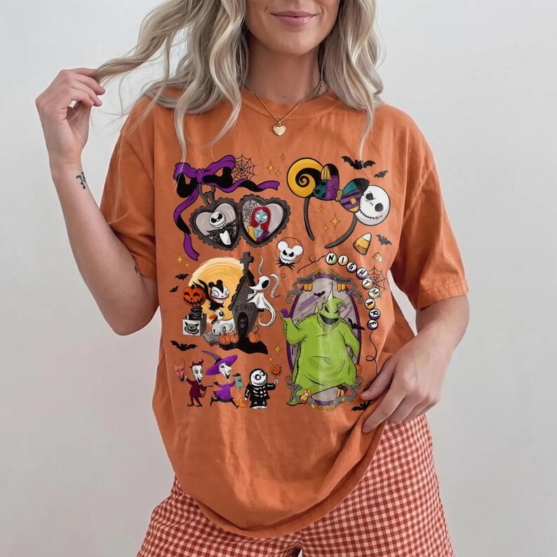 May include: Orange t-shirt with a colourful graphic design featuring characters from "The Nightmare Before Christmas." The design includes Jack Skellington, Sally, and Oogie Boogie. The shirt has a crew neck and short sleeves.