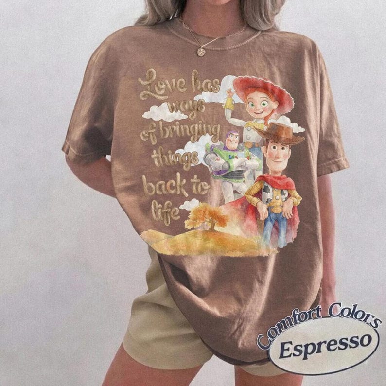 May include: A brown short-sleeve t-shirt featuring a graphic print of Toy Story characters including Woody, Buzz Lightyear, and Jessie. The text reads, Love has ways of bringing things back to life. The shirt has a crew neckline and a relaxed fit.